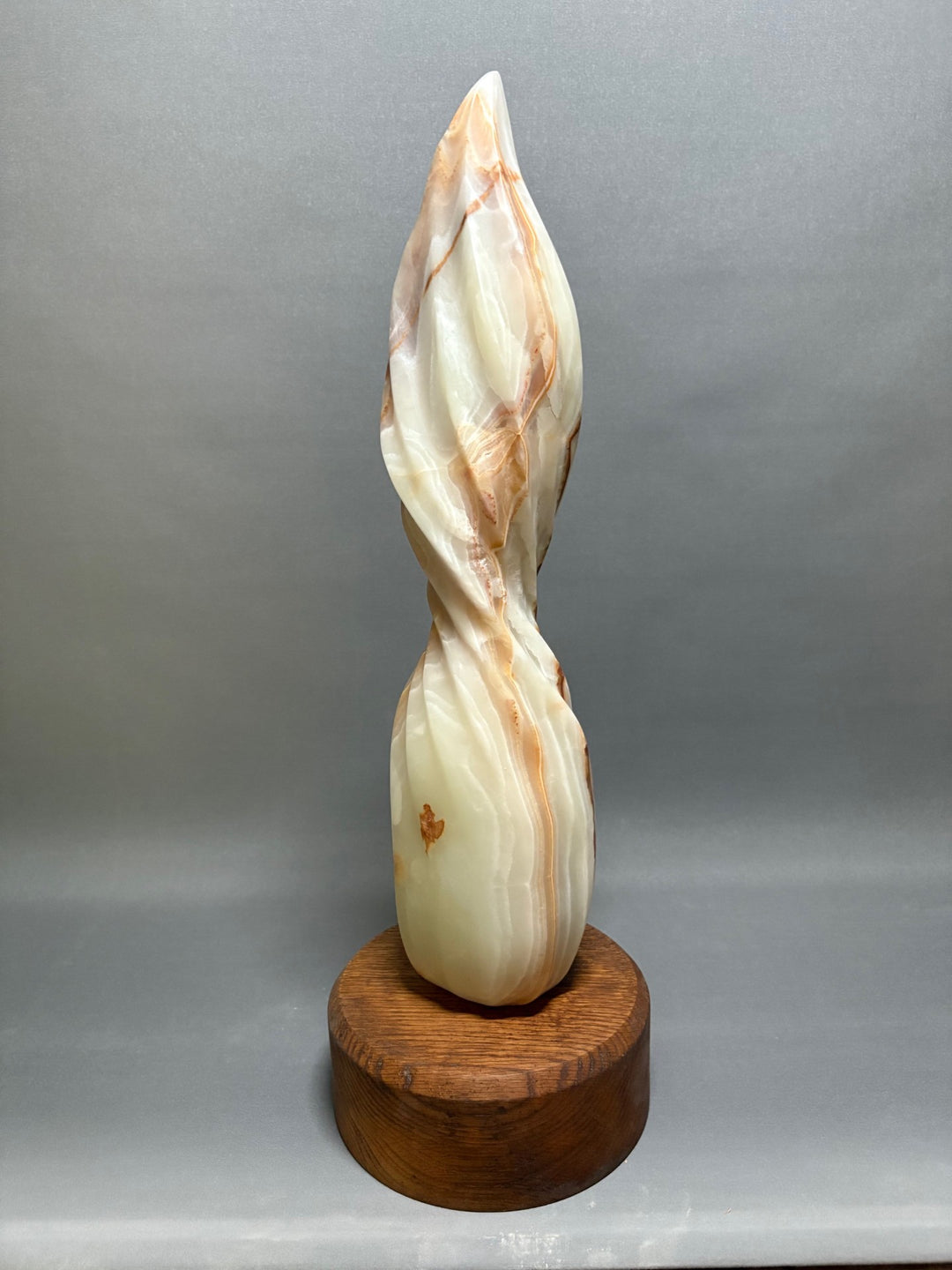 Onyx Twist by Brendan Cullen, a hand-carved Patagonian onyx sculpture with a slender, flame-like spiral form rising from a rounded wooden base, the translucent stone glowing in creamy white with warm amber and rust-coloured veining.
