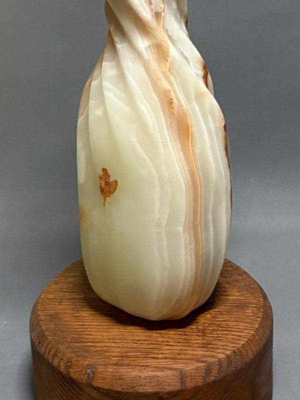 Onyx Twist by Brendan Cullen, a hand-carved Patagonian onyx sculpture with a slender, flame-like spiral form rising from a rounded wooden base, the translucent stone glowing in creamy white with warm amber and rust-coloured veining.