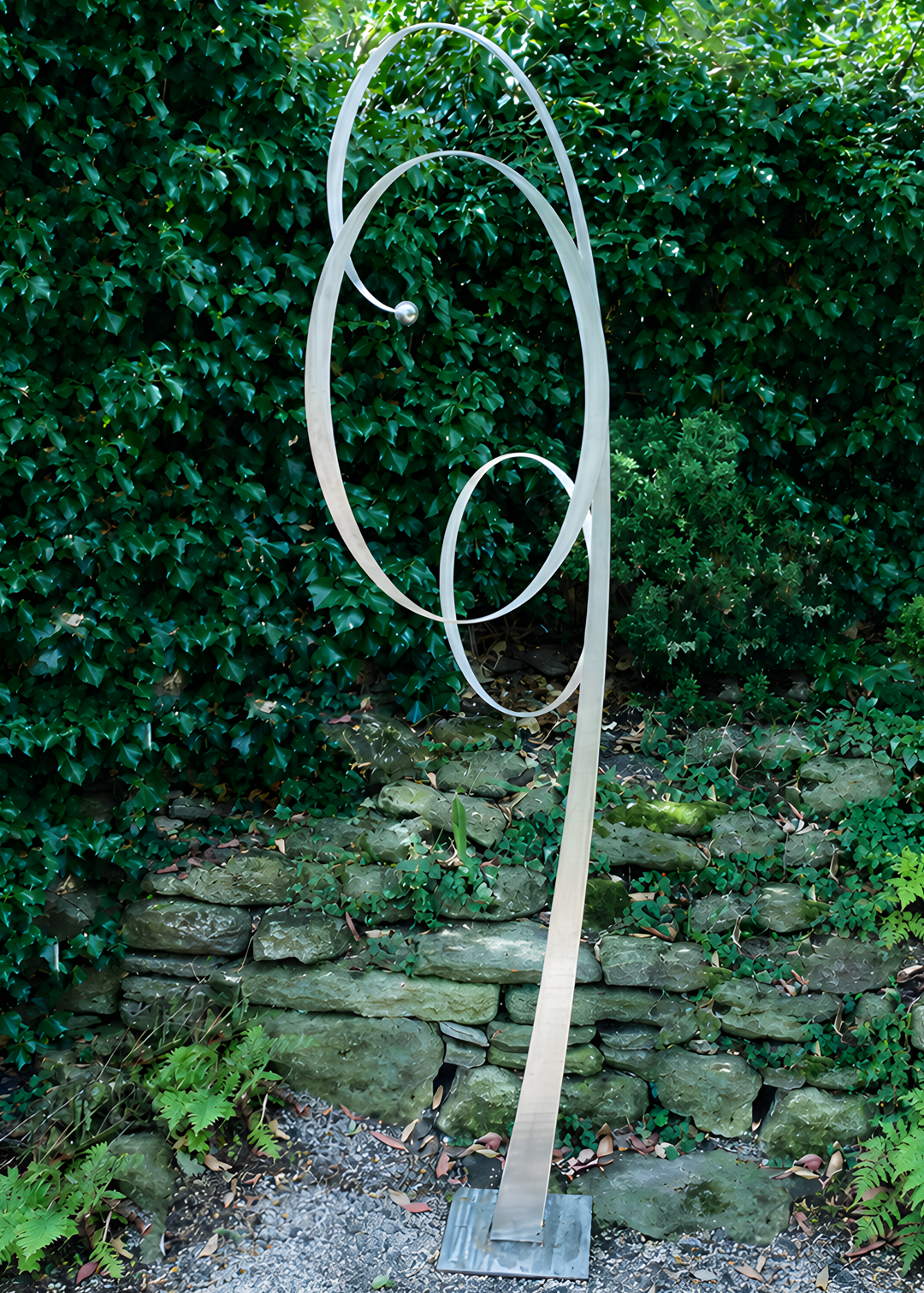 Ian Marlow MRSS stainless steel sculpture Orbiting Upwards, featuring a tall contemporary abstract form with sweeping interconnected circular loops rising upwards, displayed outdoors within a landscaped garden setting.