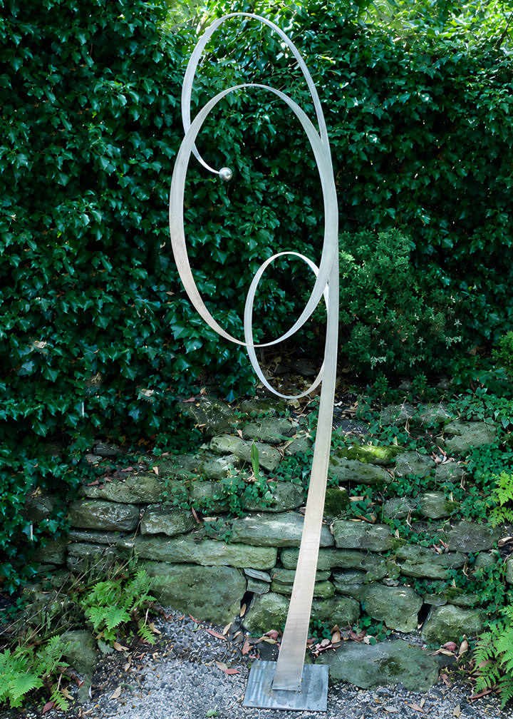 Ian Marlow MRSS stainless steel sculpture Orbiting Upwards, featuring a tall contemporary abstract form with sweeping interconnected circular loops rising upwards, displayed outdoors within a landscaped garden setting.