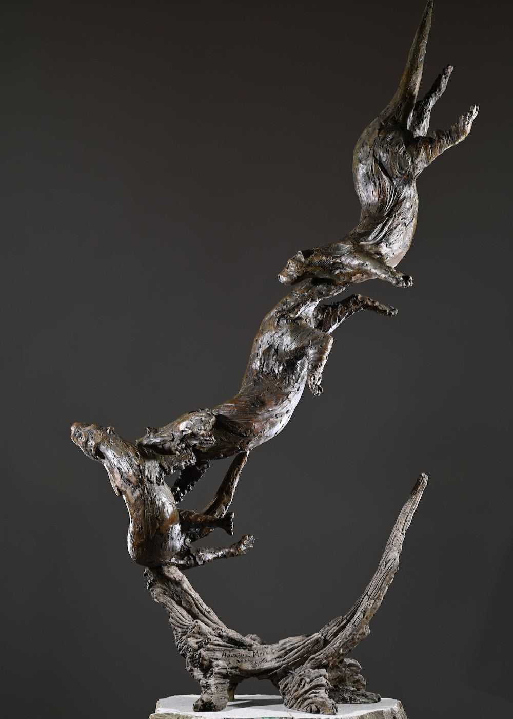 ‘Otters Playing Underwater’ by Hamish Mackie, a life-size limited-edition bronze wildlife sculpture depicting a mother otter and her playful cubs twisting, diving and swimming around a sculpted underwater tree root. The dynamic vertical composition features expressive animal movements, detailed fur textures and a weathered bronze finish in rich brown and earthy tones, capturing the playful energy and natural behaviour of otters beneath the water.