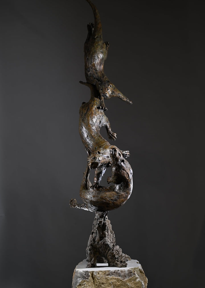 ‘Otters Playing Underwater’ by Hamish Mackie, a life-size limited-edition bronze wildlife sculpture depicting a mother otter and her playful cubs twisting, diving and swimming around a sculpted underwater tree root. The dynamic vertical composition features expressive animal movements, detailed fur textures and a weathered bronze finish in rich brown and earthy tones, capturing the playful energy and natural behaviour of otters beneath the water.