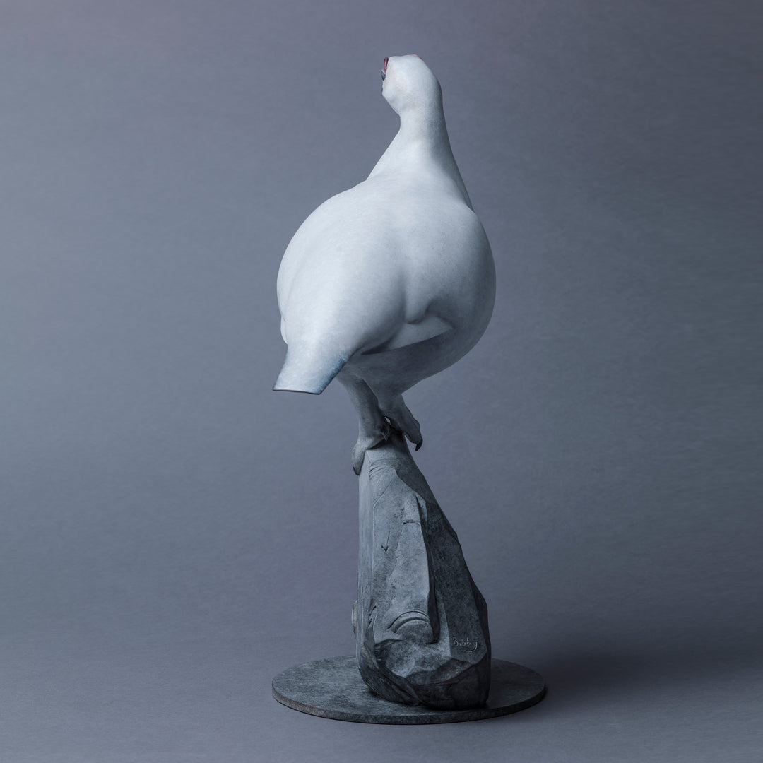 A finely sculpted Nick Bibby bronze ptarmigan standing tall on an angular rocky base, with a smooth winter-white patina and soft grey tonality, capturing the bird’s alert posture and clean, rounded form against a neutral backdrop.