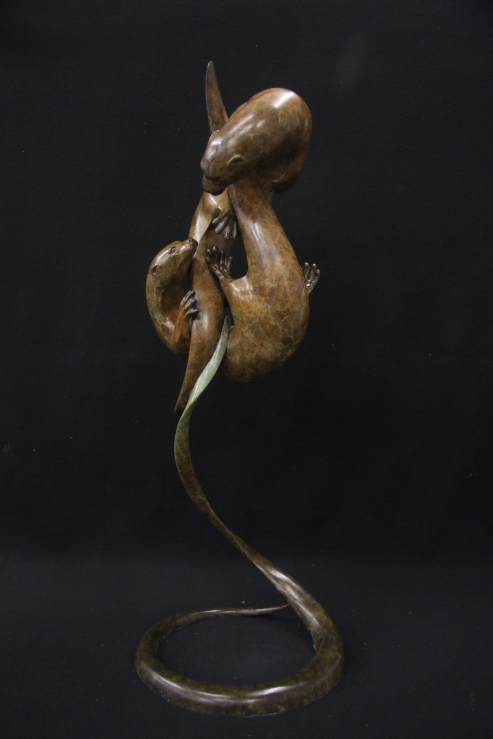 Elegant bronze sculpture of two otters playfully entwined in an upward spiral against a black background.