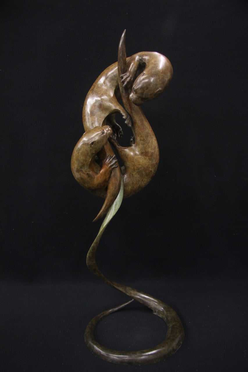 Elegant bronze sculpture of two otters playfully entwined in an upward spiral against a black background.