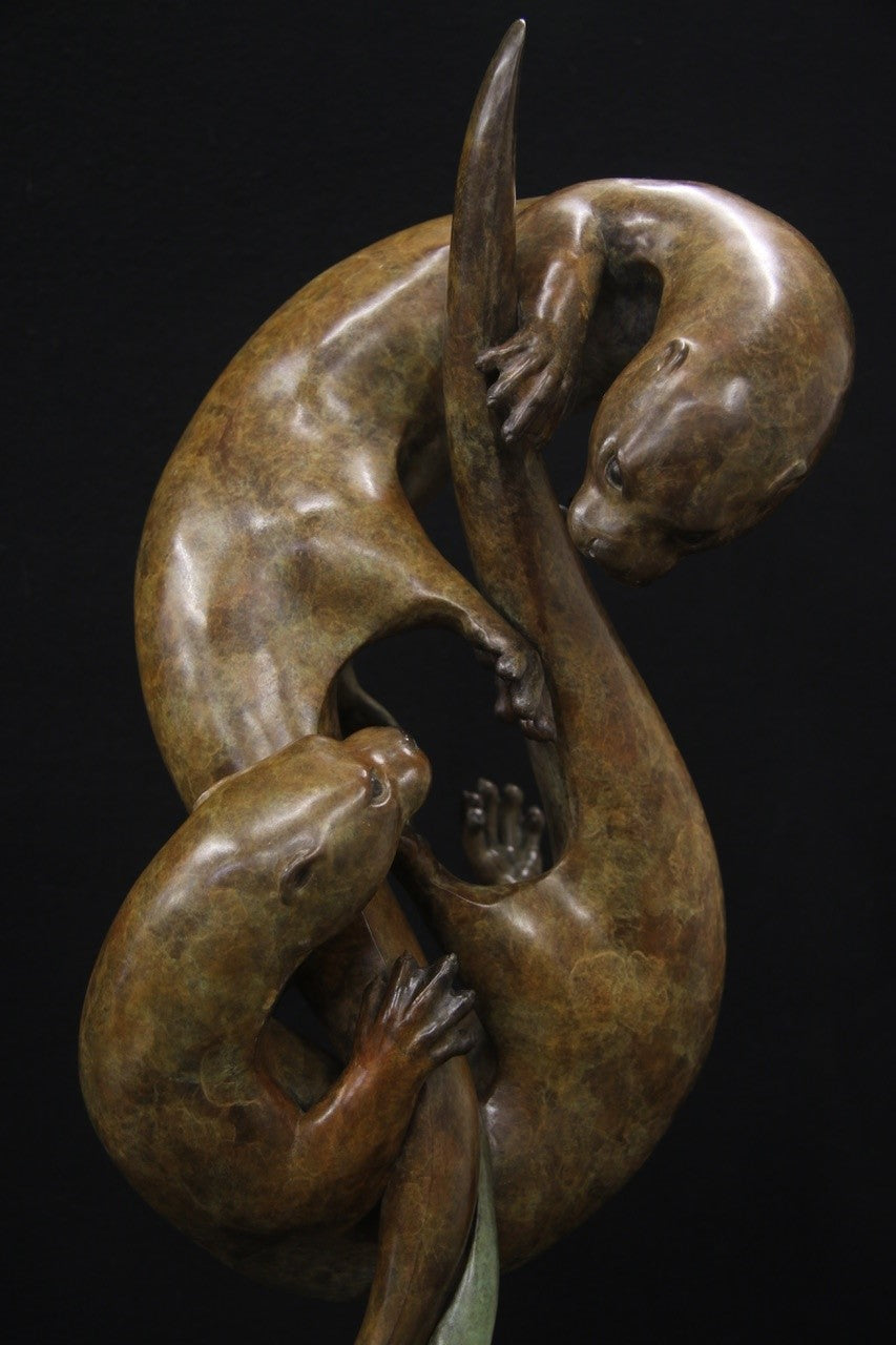 Elegant bronze sculpture of two otters playfully entwined in an upward spiral against a black background.