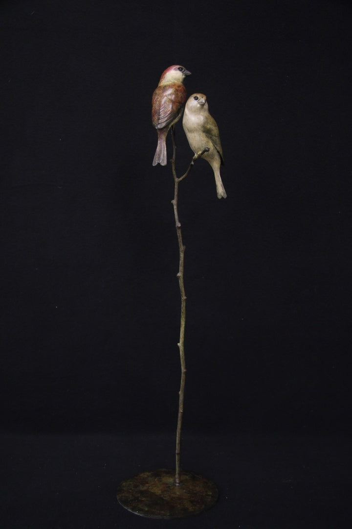 Bronze sculpture of two small sparrows birds perched closely together on a slender, vertical branch with a circular base, set against a black background.