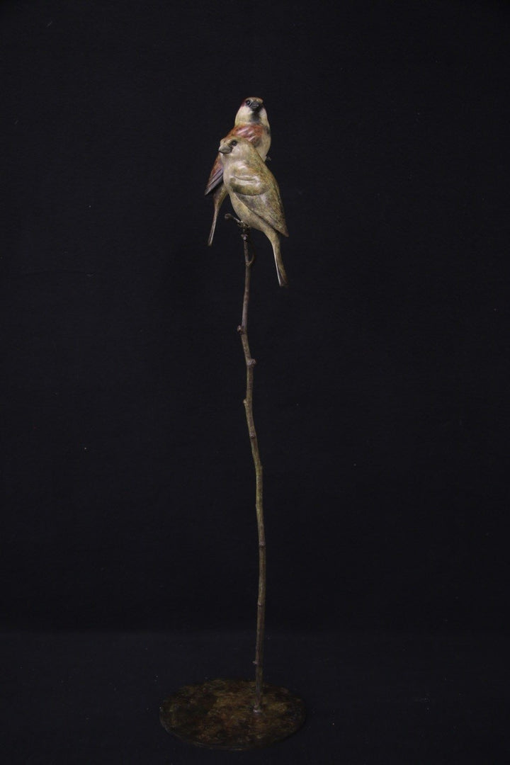 Bronze sculpture of two small sparrows birds perched closely together on a slender, vertical branch with a circular base, set against a black background.
