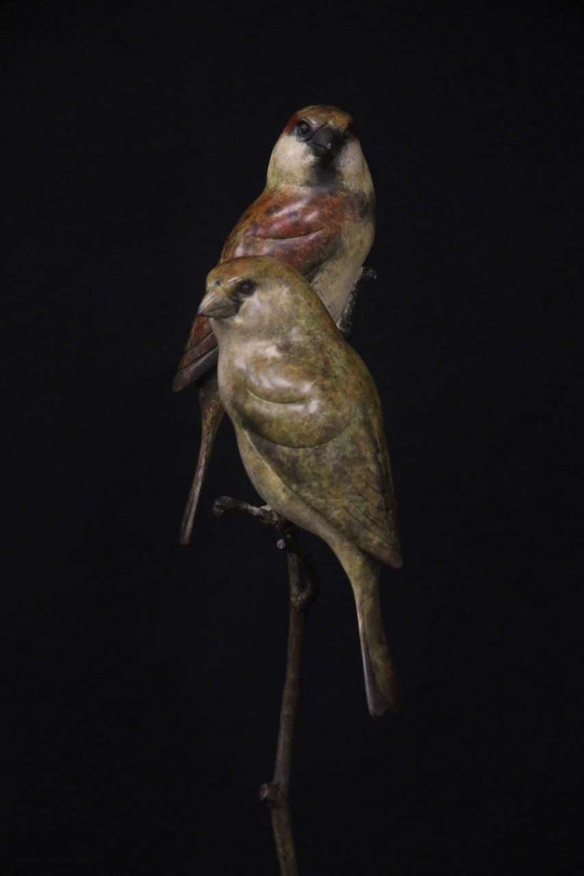 Bronze sculpture of two small sparrows birds perched closely together on a slender, vertical branch with a circular base, set against a black background.