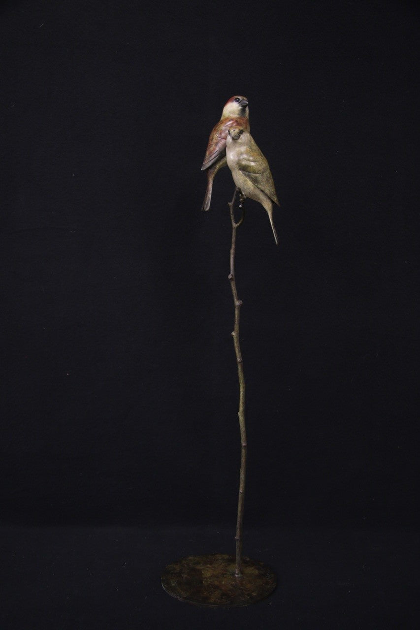 Bronze sculpture of two small sparrows birds perched closely together on a slender, vertical branch with a circular base, set against a black background.