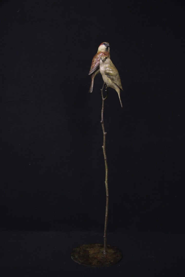 Bronze sculpture of two small sparrows birds perched closely together on a slender, vertical branch with a circular base, set against a black background.