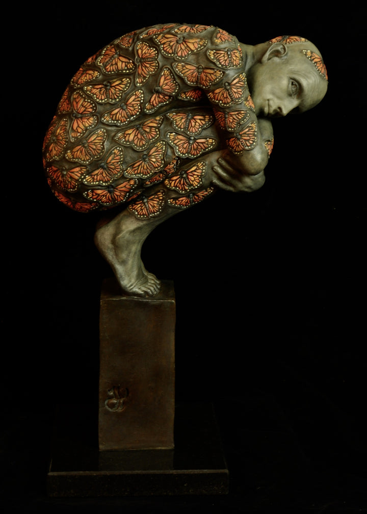 Bronze sculpture of a crouched male figure balanced on a square plinth, his body wrapped tightly around itself in a contemplative pose. The entire surface is patterned with vivid monarch butterflies in orange, black and soft yellow tones, creating a striking contrast against the subdued bronze of the skin. His expression is introspective and hesitant, poised between stillness and the potential for movement. Set against a dark background, the sculpture highlights themes of transformation and courage.