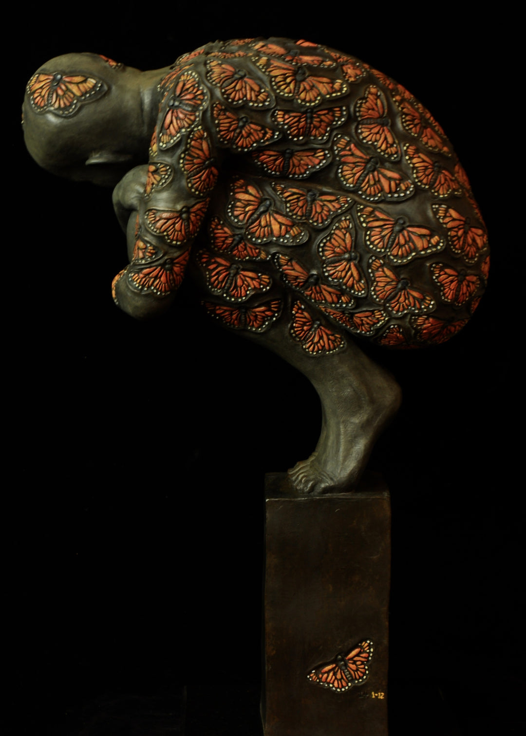 Bronze sculpture of a crouched male figure balanced on a square plinth, his body wrapped tightly around itself in a contemplative pose. The entire surface is patterned with vivid monarch butterflies in orange, black and soft yellow tones, creating a striking contrast against the subdued bronze of the skin. His expression is introspective and hesitant, poised between stillness and the potential for movement. Set against a dark background, the sculpture highlights themes of transformation and courage.