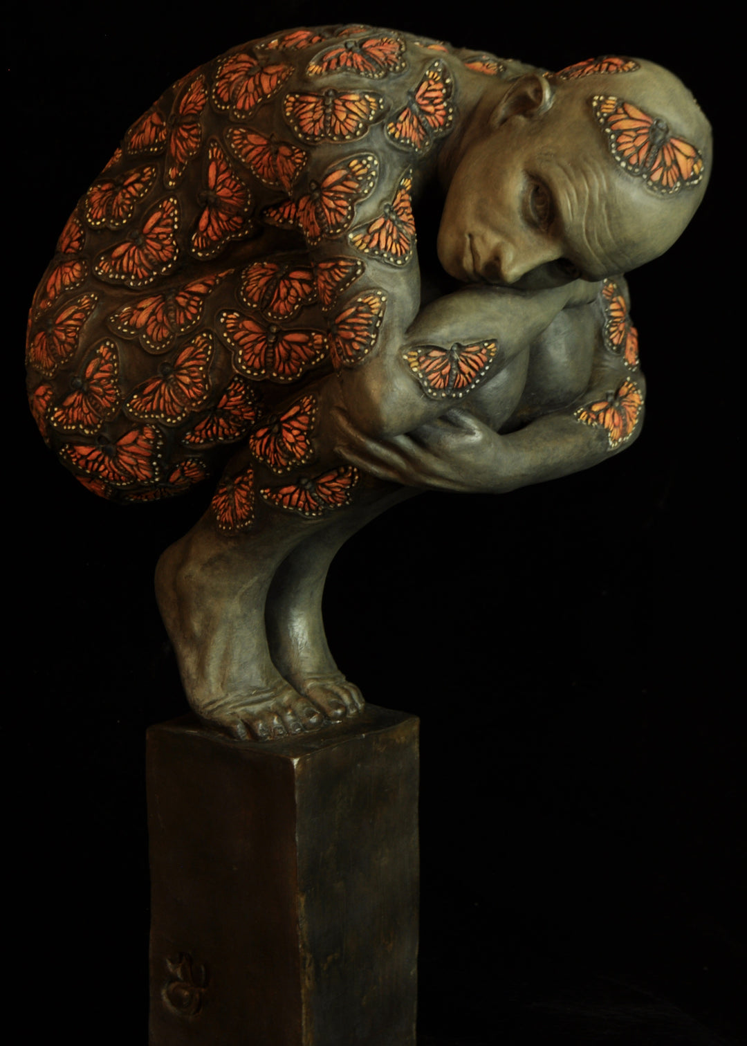 Bronze sculpture of a crouched male figure balanced on a square plinth, his body wrapped tightly around itself in a contemplative pose. The entire surface is patterned with vivid monarch butterflies in orange, black and soft yellow tones, creating a striking contrast against the subdued bronze of the skin. His expression is introspective and hesitant, poised between stillness and the potential for movement. Set against a dark background, the sculpture highlights themes of transformation and courage.