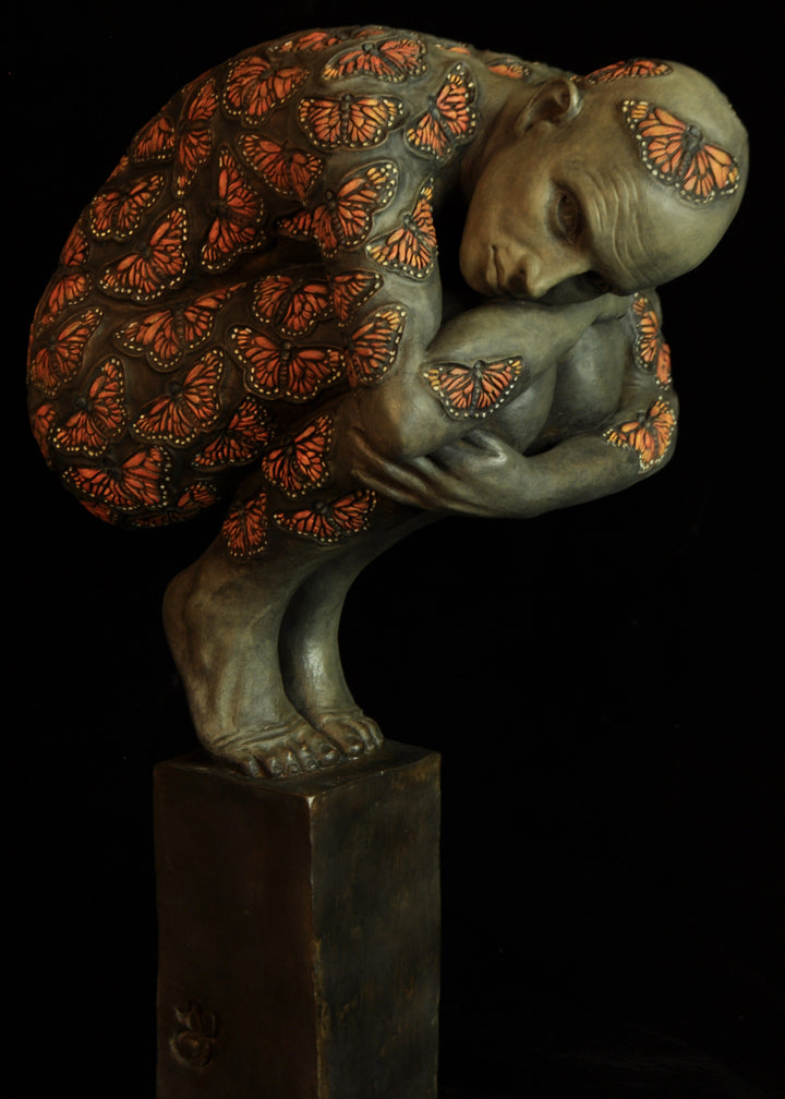 Bronze sculpture of a crouched male figure balanced on a square plinth, his body wrapped tightly around itself in a contemplative pose. The entire surface is patterned with vivid monarch butterflies in orange, black and soft yellow tones, creating a striking contrast against the subdued bronze of the skin. His expression is introspective and hesitant, poised between stillness and the potential for movement. Set against a dark background, the sculpture highlights themes of transformation and courage.