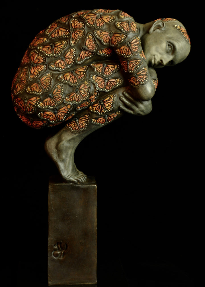 Bronze sculpture of a crouched male figure balanced on a square plinth, his body wrapped tightly around itself in a contemplative pose. The entire surface is patterned with vivid monarch butterflies in orange, black and soft yellow tones, creating a striking contrast against the subdued bronze of the skin. His expression is introspective and hesitant, poised between stillness and the potential for movement. Set against a dark background, the sculpture highlights themes of transformation and courage.