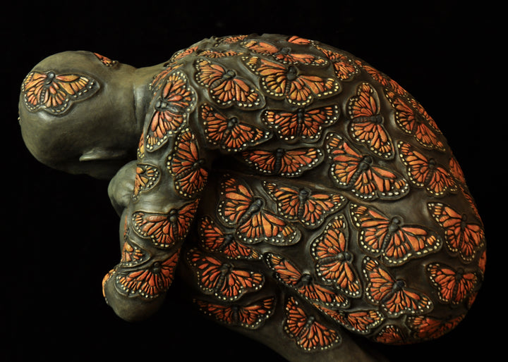Bronze sculpture of a crouched male figure balanced on a square plinth, his body wrapped tightly around itself in a contemplative pose. The entire surface is patterned with vivid monarch butterflies in orange, black and soft yellow tones, creating a striking contrast against the subdued bronze of the skin. His expression is introspective and hesitant, poised between stillness and the potential for movement. Set against a dark background, the sculpture highlights themes of transformation and courage.