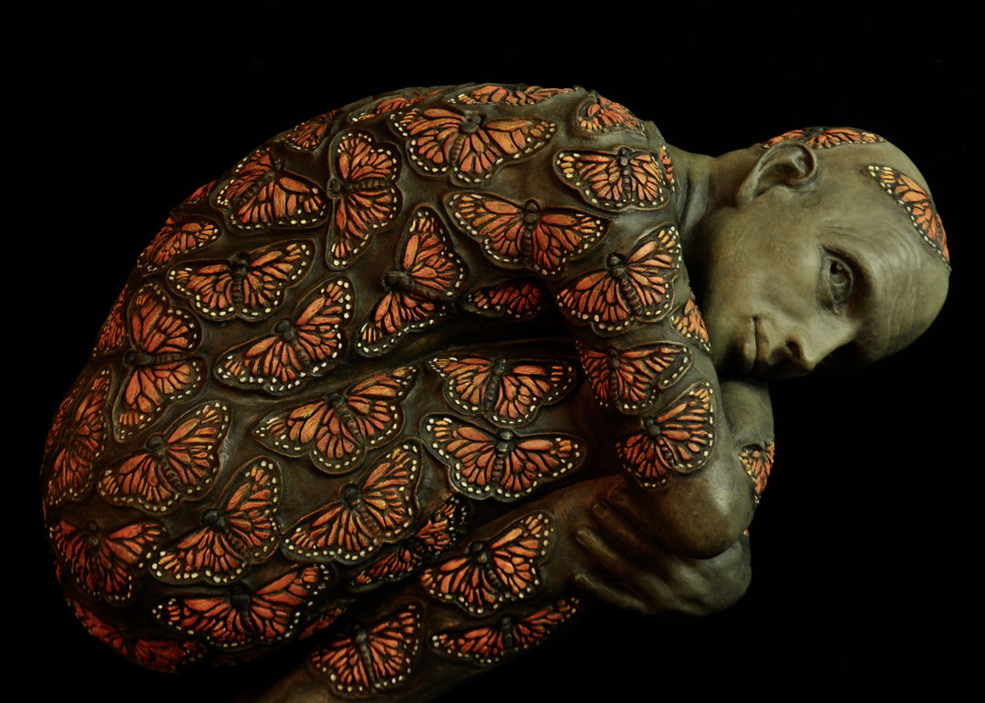 Bronze sculpture of a crouched male figure balanced on a square plinth, his body wrapped tightly around itself in a contemplative pose. The entire surface is patterned with vivid monarch butterflies in orange, black and soft yellow tones, creating a striking contrast against the subdued bronze of the skin. His expression is introspective and hesitant, poised between stillness and the potential for movement. Set against a dark background, the sculpture highlights themes of transformation and courage.