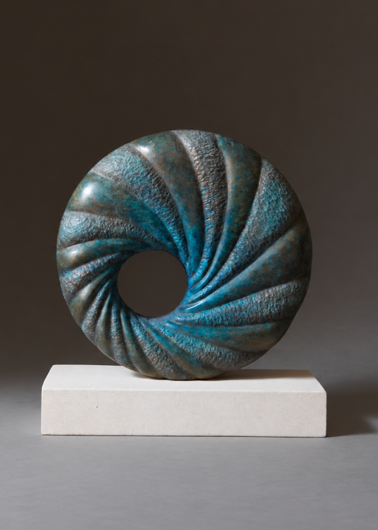 ‘Praa’ by Nicolas Moreton MRSS, a limited-edition contemporary abstract bronze sculpture featuring a circular form with a central opening and deeply carved spiralling ridges. Alternating smooth and textured surfaces are finished in turquoise, teal, blue-green and dark bronze patina, creating a twisting pattern around the open centre. The sculpture is displayed on a rectangular pale cream stone base against a dark grey and brown studio background.