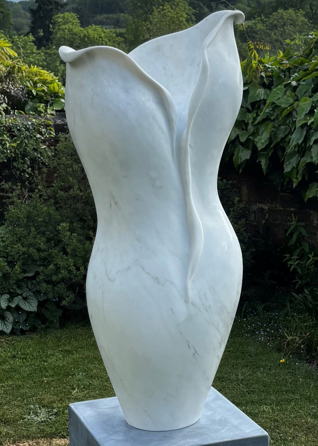 Elegant outdoor marble sculpture titled “Petali” by Lucy Unwin, hand-carved from luminous white Carrara marble with delicate petal-like curves and fluid organic lines, standing gracefully on a smooth stone plinth against a natural garden backdrop.