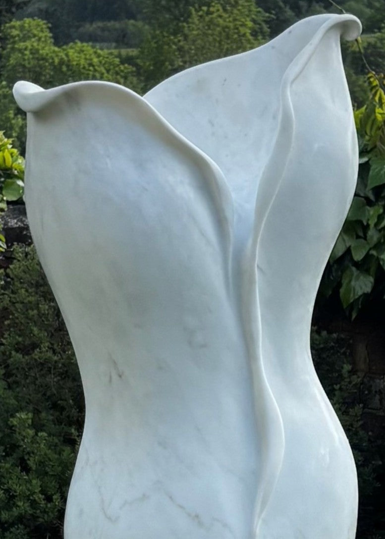 Elegant outdoor marble sculpture titled “Petali” by Lucy Unwin, hand-carved from luminous white Carrara marble with delicate petal-like curves and fluid organic lines, standing gracefully on a smooth stone plinth against a natural garden backdrop.