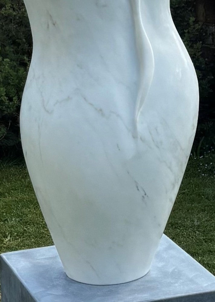 Elegant outdoor marble sculpture titled “Petali” by Lucy Unwin, hand-carved from luminous white Carrara marble with delicate petal-like curves and fluid organic lines, standing gracefully on a smooth stone plinth against a natural garden backdrop.