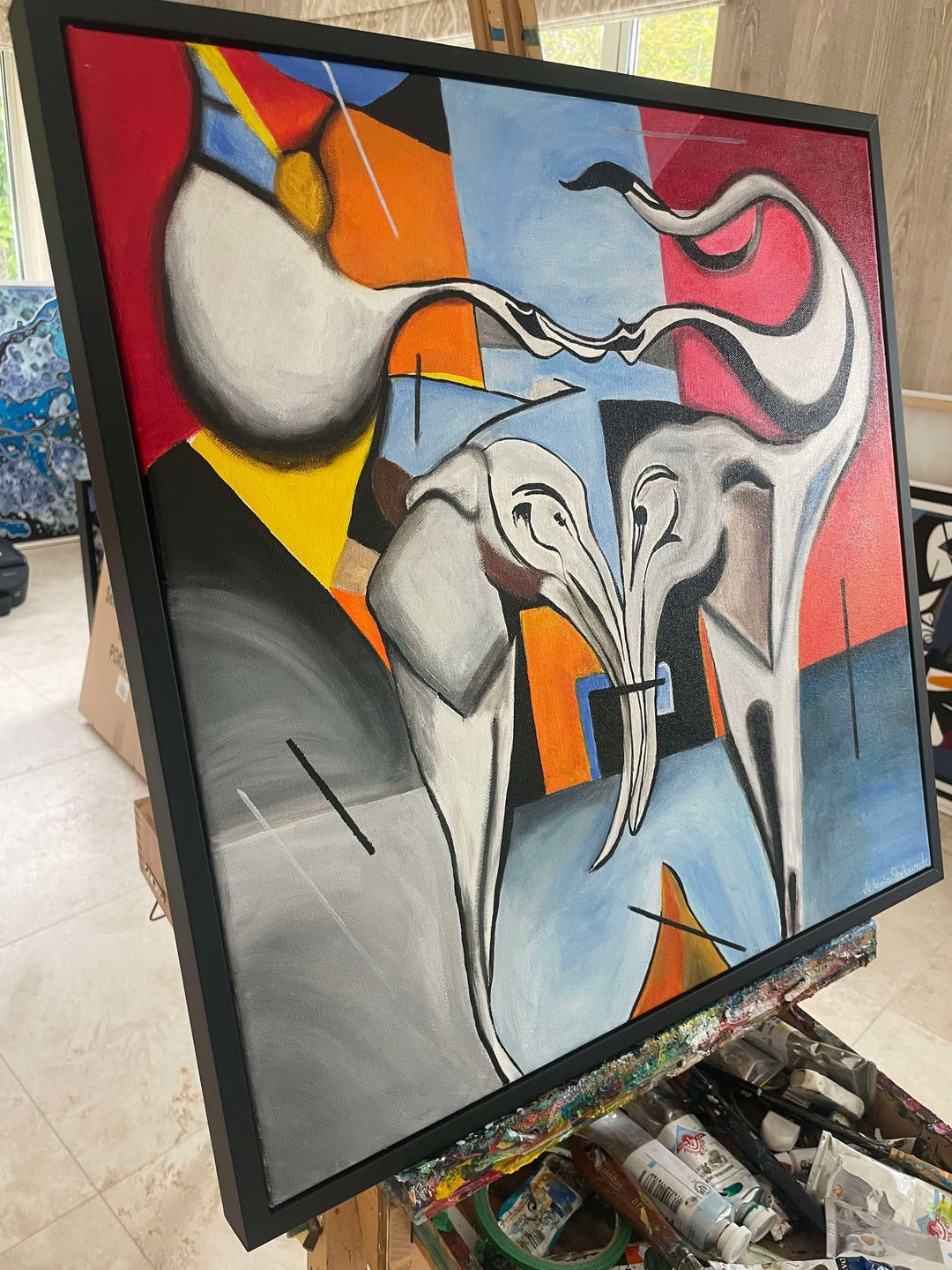 Abstract painting of two stylised elephants with entwined trunks, set against a bold geometric background in red, blue, yellow, and black.

