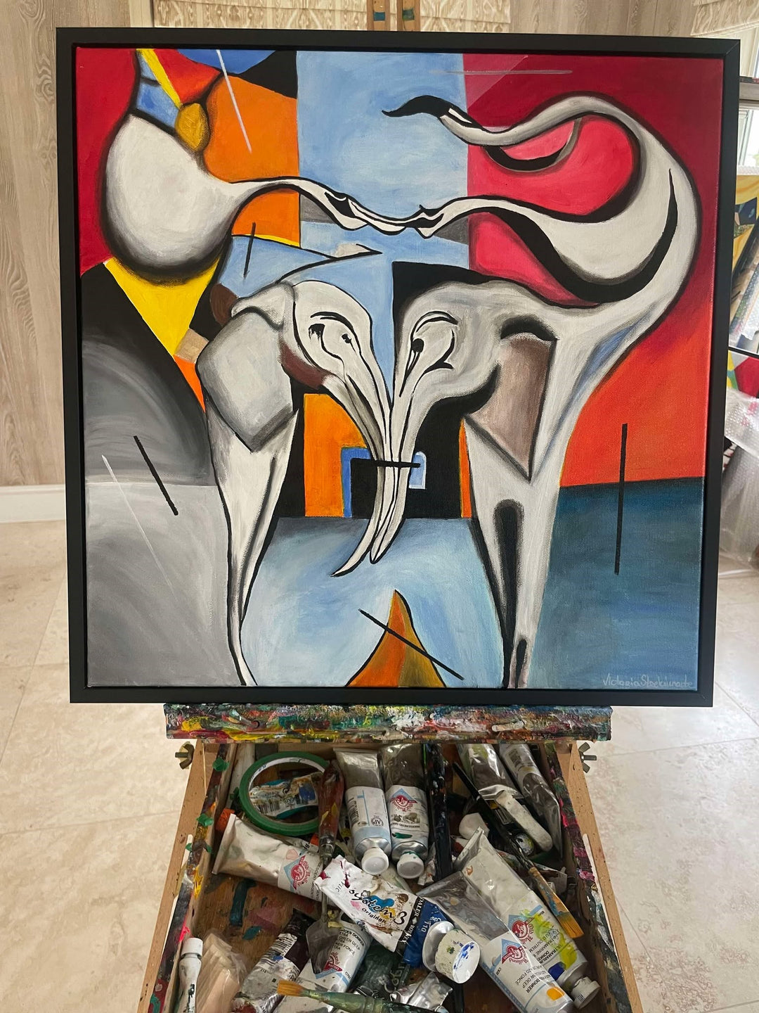 Abstract painting of two stylised elephants with entwined trunks, set against a bold geometric background in red, blue, yellow, and black.

