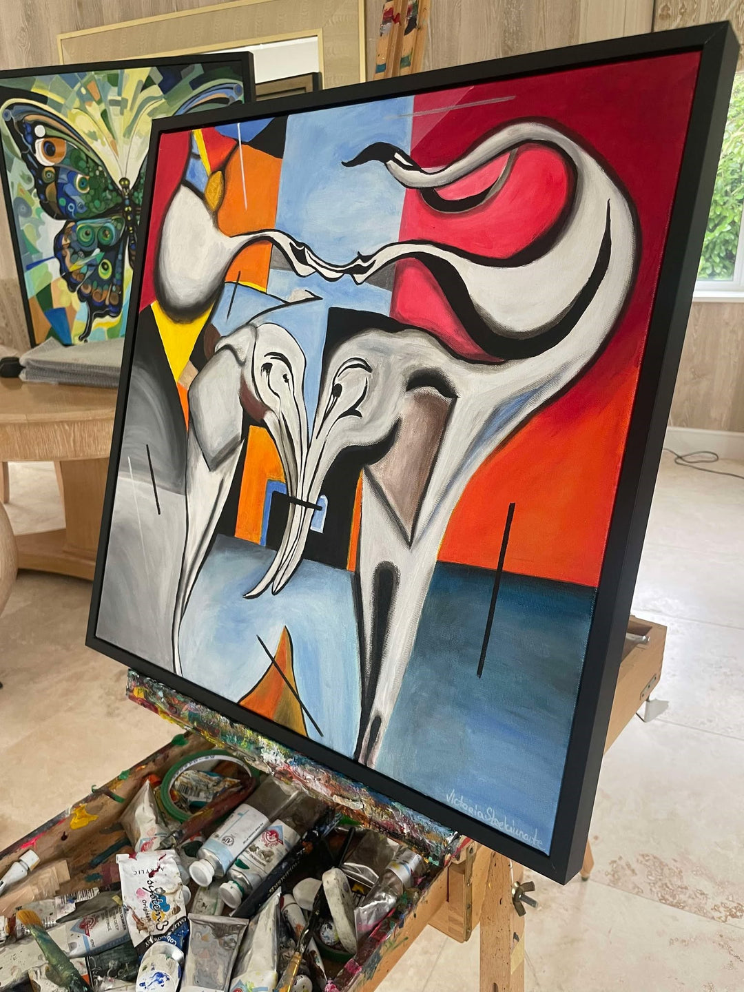 Abstract painting of two stylised elephants with entwined trunks, set against a bold geometric background in red, blue, yellow, and black.

