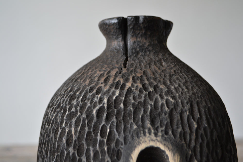 Tall, vertical abstract sculpture by Nicola Beattie, meticulously hand-carved from scorched sycamore. The monolithic form features a deep, elongated central hollow framed by natural wood highlights against a richly textured, blackened surface. Mounted on a solid square base, the piece evokes ancient standing stones and architectural ruins, with a tactile finish that invites close inspection. Its black and warm beige tones, organic curves, and minimalist presence make it a striking statement for interiors.