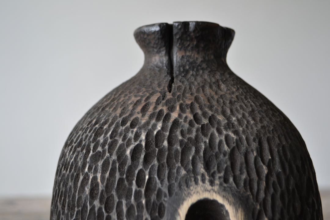 Tall, vertical abstract sculpture by Nicola Beattie, meticulously hand-carved from scorched sycamore. The monolithic form features a deep, elongated central hollow framed by natural wood highlights against a richly textured, blackened surface. Mounted on a solid square base, the piece evokes ancient standing stones and architectural ruins, with a tactile finish that invites close inspection. Its black and warm beige tones, organic curves, and minimalist presence make it a striking statement for interiors.