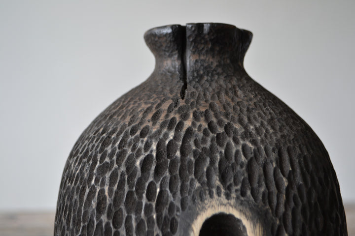 Tall, vertical abstract sculpture by Nicola Beattie, meticulously hand-carved from scorched sycamore. The monolithic form features a deep, elongated central hollow framed by natural wood highlights against a richly textured, blackened surface. Mounted on a solid square base, the piece evokes ancient standing stones and architectural ruins, with a tactile finish that invites close inspection. Its black and warm beige tones, organic curves, and minimalist presence make it a striking statement for interiors.