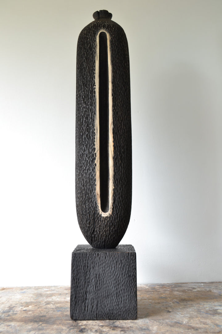 Tall, vertical abstract sculpture by Nicola Beattie, meticulously hand-carved from scorched sycamore. The monolithic form features a deep, elongated central hollow framed by natural wood highlights against a richly textured, blackened surface. Mounted on a solid square base, the piece evokes ancient standing stones and architectural ruins, with a tactile finish that invites close inspection. Its black and warm beige tones, organic curves, and minimalist presence make it a striking statement for interiors.