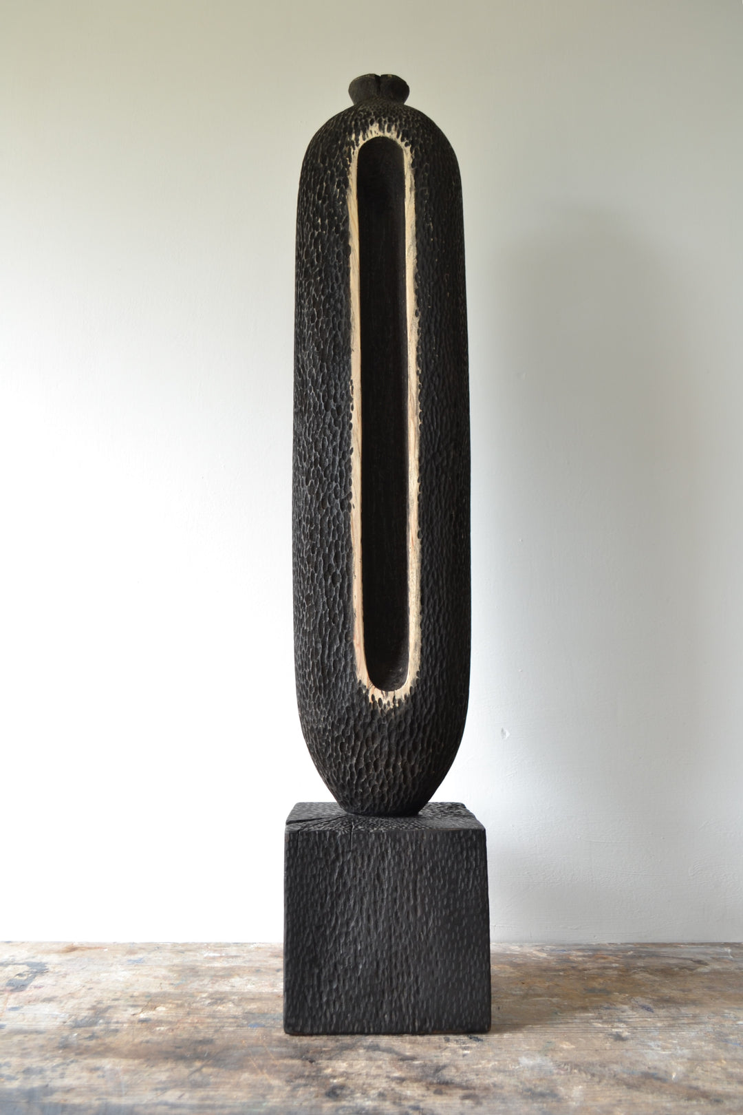Tall, vertical abstract sculpture by Nicola Beattie, meticulously hand-carved from scorched sycamore. The monolithic form features a deep, elongated central hollow framed by natural wood highlights against a richly textured, blackened surface. Mounted on a solid square base, the piece evokes ancient standing stones and architectural ruins, with a tactile finish that invites close inspection. Its black and warm beige tones, organic curves, and minimalist presence make it a striking statement for interiors.