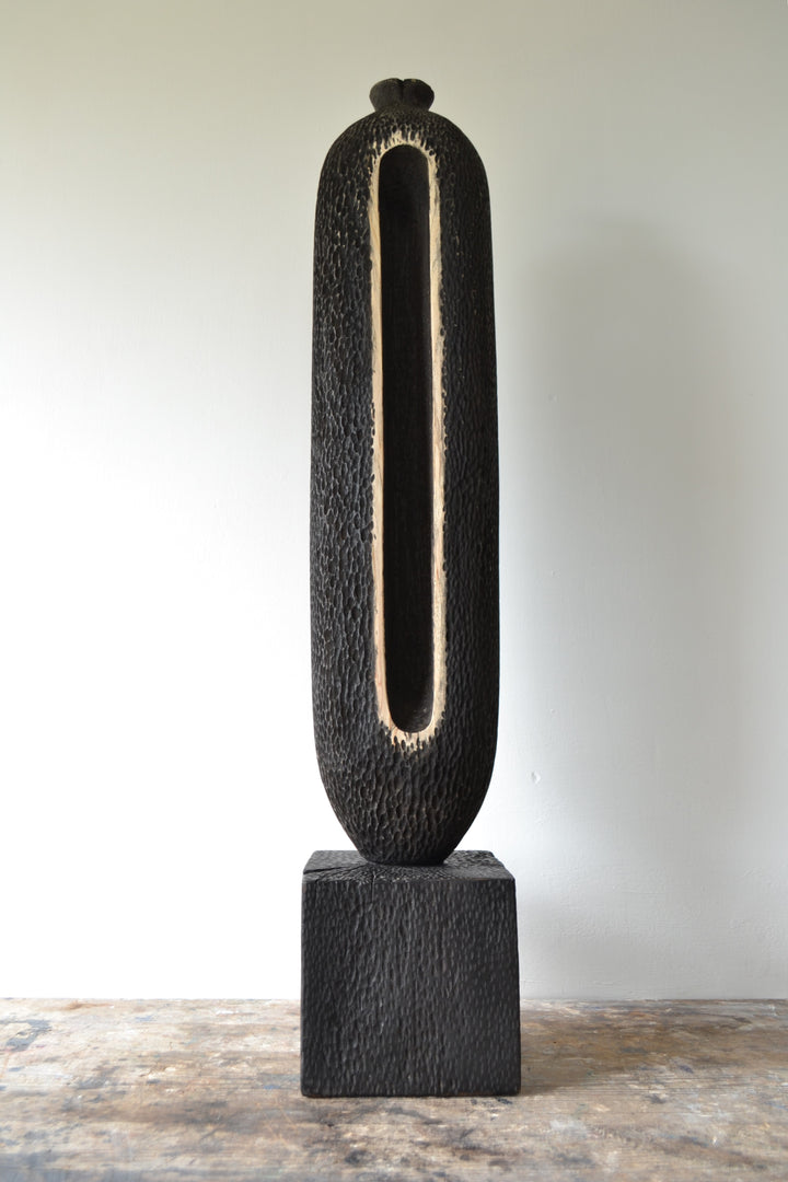 Tall, vertical abstract sculpture by Nicola Beattie, meticulously hand-carved from scorched sycamore. The monolithic form features a deep, elongated central hollow framed by natural wood highlights against a richly textured, blackened surface. Mounted on a solid square base, the piece evokes ancient standing stones and architectural ruins, with a tactile finish that invites close inspection. Its black and warm beige tones, organic curves, and minimalist presence make it a striking statement for interiors.