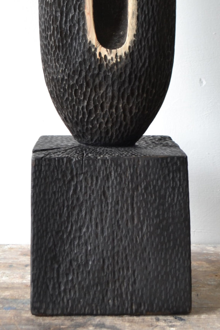Tall, vertical abstract sculpture by Nicola Beattie, meticulously hand-carved from scorched sycamore. The monolithic form features a deep, elongated central hollow framed by natural wood highlights against a richly textured, blackened surface. Mounted on a solid square base, the piece evokes ancient standing stones and architectural ruins, with a tactile finish that invites close inspection. Its black and warm beige tones, organic curves, and minimalist presence make it a striking statement for interiors.