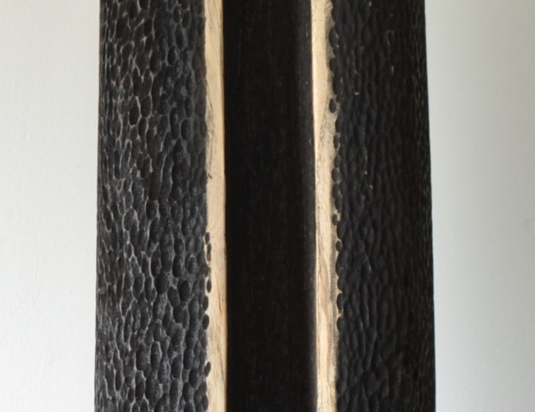 Tall, vertical abstract sculpture by Nicola Beattie, meticulously hand-carved from scorched sycamore. The monolithic form features a deep, elongated central hollow framed by natural wood highlights against a richly textured, blackened surface. Mounted on a solid square base, the piece evokes ancient standing stones and architectural ruins, with a tactile finish that invites close inspection. Its black and warm beige tones, organic curves, and minimalist presence make it a striking statement for interiors.