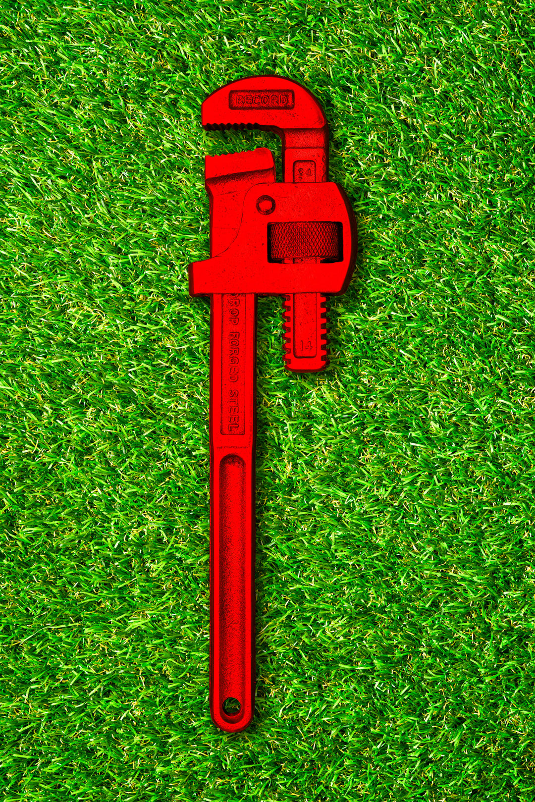 Photograph of a bold red pipe wrench laid flat on vibrant green grass, contrasting industrial form with natural texture.