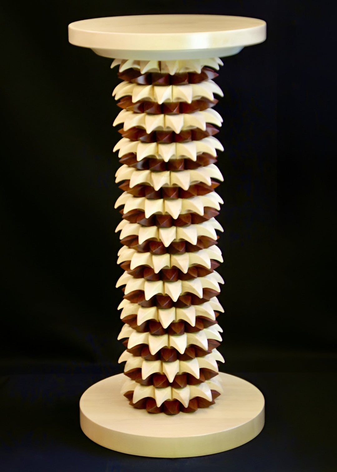 Sculptural table in maple and recycled mahogany, with a cylindrical base of layered pollen-like geometric forms in pale wood and deep red-brown, topped with a smooth circular surface.