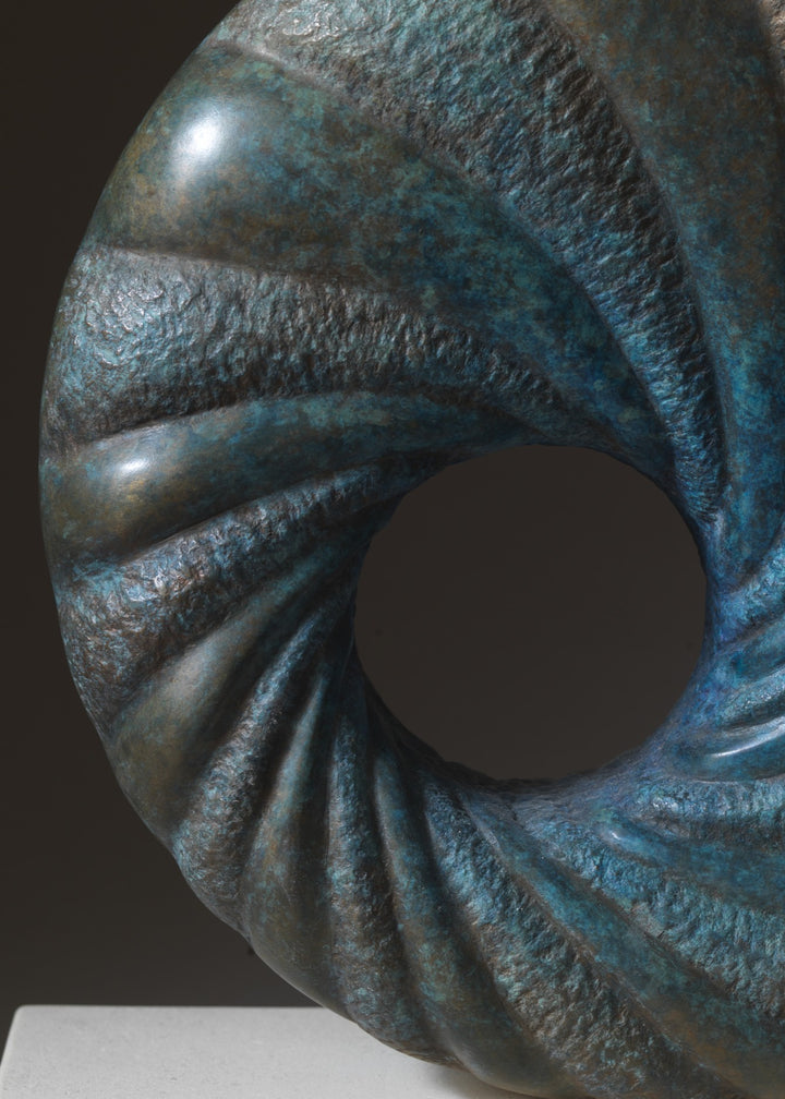 ‘Praa’ by Nicolas Moreton MRSS, a limited-edition contemporary abstract bronze sculpture featuring a circular form with a central opening and deeply carved spiralling ridges. Alternating smooth and textured surfaces are finished in turquoise, teal, blue-green and dark bronze patina, creating a twisting pattern around the open centre. The sculpture is displayed on a rectangular pale cream stone base against a dark grey and brown studio background.