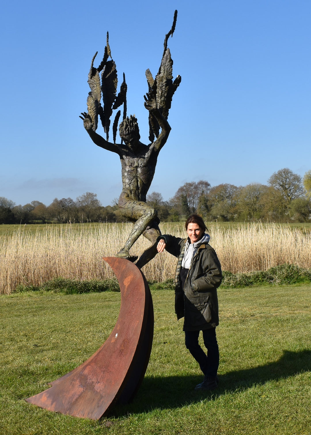 Monumental outdoor bronze sculpture by Nicola Godden — a dramatic, winged male figure with raised arms, climbing and balancing on a sweeping rusted steel crescent. The figure’s body has a textured verdigris green and earthy bronze patina with highlights of gold and brown, contrasting with the deep weathered red-orange steel base. Photographed in an open grassy field with blue sky.