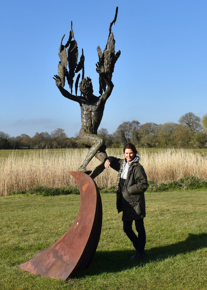 Monumental outdoor bronze sculpture by Nicola Godden — a dramatic, winged male figure with raised arms, climbing and balancing on a sweeping rusted steel crescent. The figure’s body has a textured verdigris green and earthy bronze patina with highlights of gold and brown, contrasting with the deep weathered red-orange steel base. Photographed in an open grassy field with blue sky.