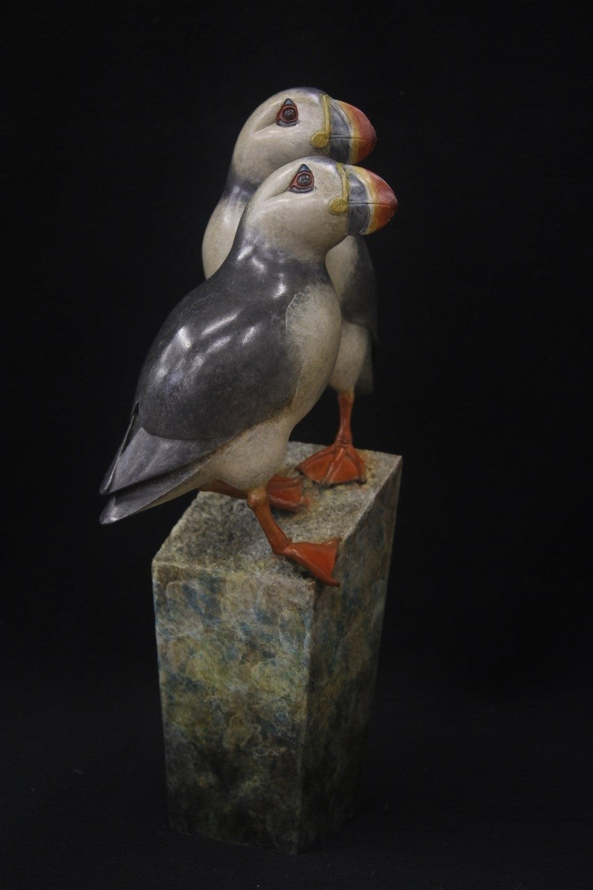 Bronze sculpture of two puffins standing side by side on a tall, textured stone plinth, featuring intricate detailing and vivid colour accents.