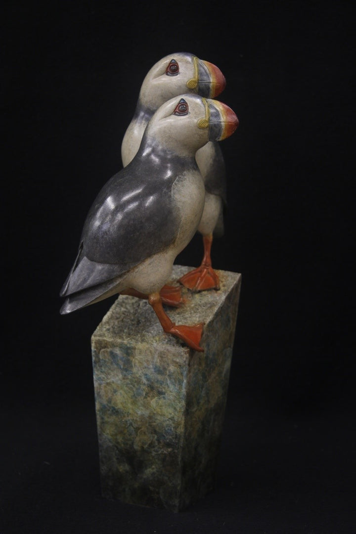 Bronze sculpture of two puffins standing side by side on a tall, textured stone plinth, featuring intricate detailing and vivid colour accents.