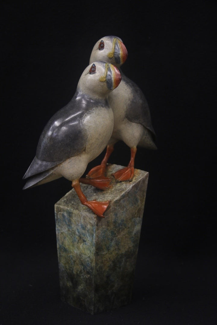 Bronze sculpture of two puffins standing side by side on a tall, textured stone plinth, featuring intricate detailing and vivid colour accents.