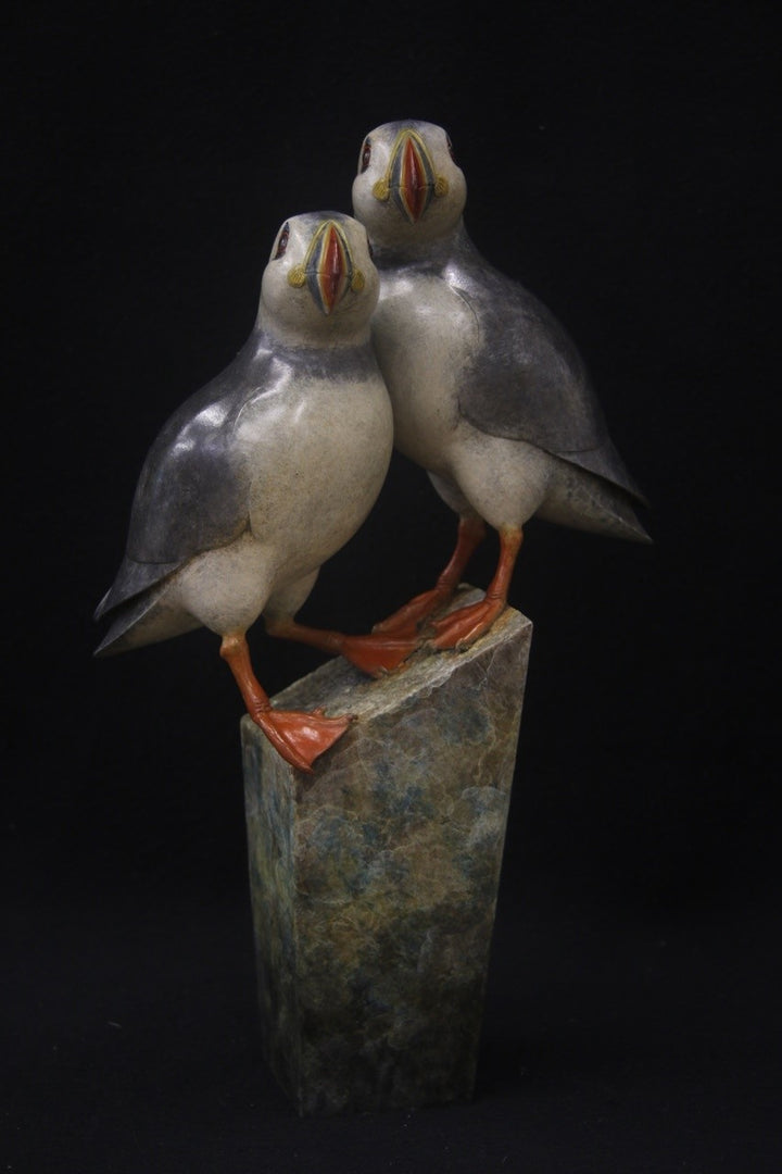 Bronze sculpture of two puffins standing side by side on a tall, textured stone plinth, featuring intricate detailing and vivid colour accents.