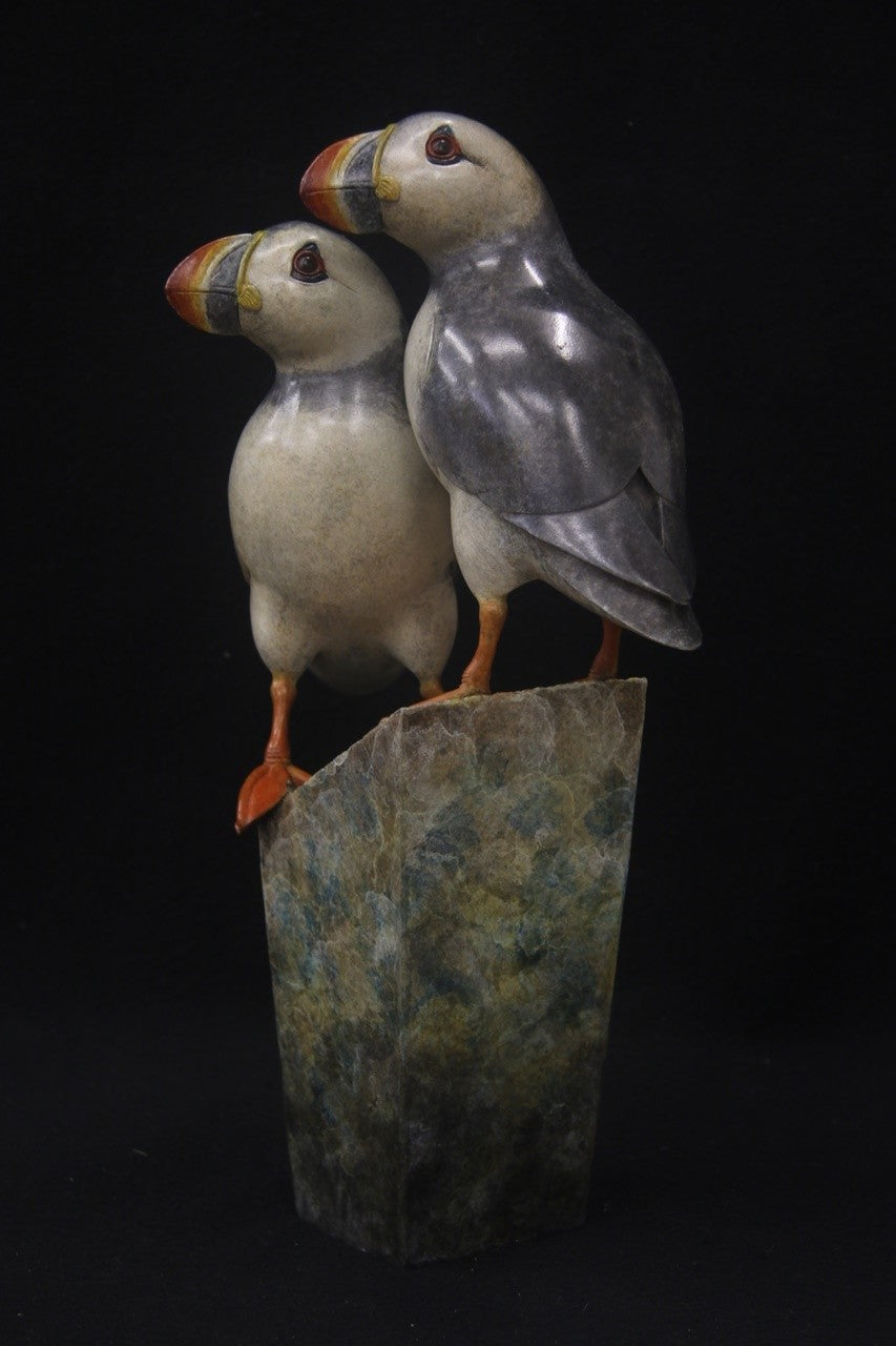 Bronze sculpture of two puffins standing side by side on a tall, textured stone plinth, featuring intricate detailing and vivid colour accents.