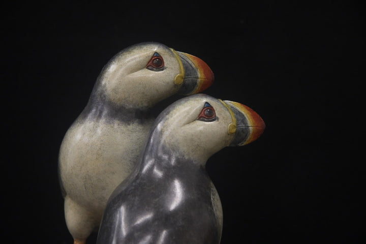 Bronze sculpture of two puffins standing side by side on a tall, textured stone plinth, featuring intricate detailing and vivid colour accents.