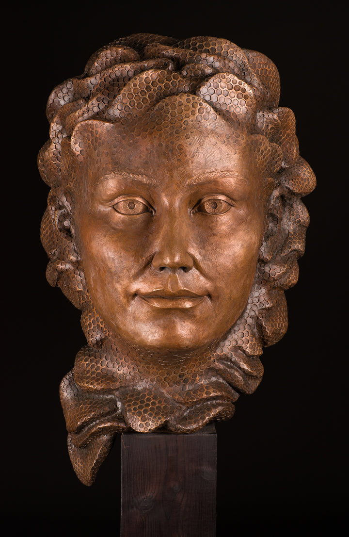Detailed bronze sculpture of a serene female face emerging from a swirling form of honeycomb texture, symbolising a queen bee. The head is crowned with hexagonal cells, flowing seamlessly into a coiled, hive-like base. The surface glows with rich golden and amber tones, celebrating fertility, nature, and the intricate structure of the hive. A bold fusion of natural pattern and human portraiture that highlights the interdependence of bees and humans.