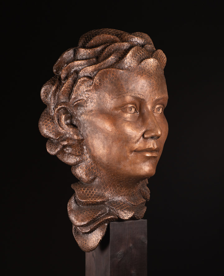 Detailed bronze sculpture of a serene female face emerging from a swirling form of honeycomb texture, symbolising a queen bee. The head is crowned with hexagonal cells, flowing seamlessly into a coiled, hive-like base. The surface glows with rich golden and amber tones, celebrating fertility, nature, and the intricate structure of the hive. A bold fusion of natural pattern and human portraiture that highlights the interdependence of bees and humans.